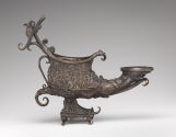 Alternate view of a bronze lamp that is shaped like a shoe, and rests on a pyramidal base.  It …