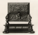 Bronze casket with protruding head in the middle at the back and various intricate motifs throu…