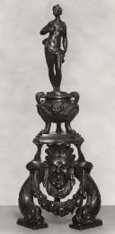 A bronze firedog with a figure of Venus.  She has her right hand on her right breast with a bag…