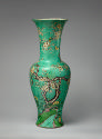 Tall porcelain vase with green ground and floral and vegetal designs on base against wood panel…