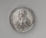 Silver medal of a woman in profile to the left wearing earrings, pearl strands woven throughout…