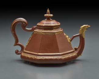 Red stoneware teapot with cover.  There are gilt details at the spout and along the top, bottom…