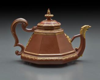 Red stoneware teapot with cover.  There are gilt details at the spout and along the top, bottom…