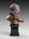 Back view of a terracotta sculpture of a head of an angel.  The angel has curly hair, which gat…