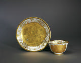 View of upright saucer with an entirely gilded chinoiserie scene in center and accompanying tea…